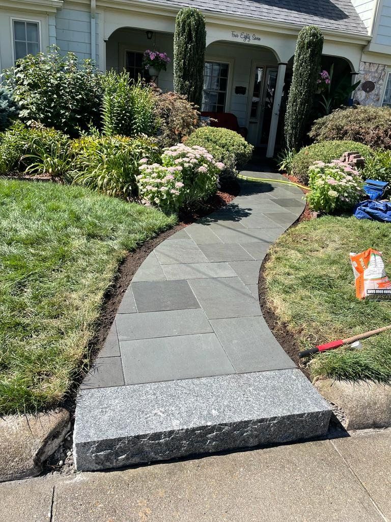 A new bluestone walkway leads to a house entrance, featuring a wide granite step at the start and landscaped bushes nearby.