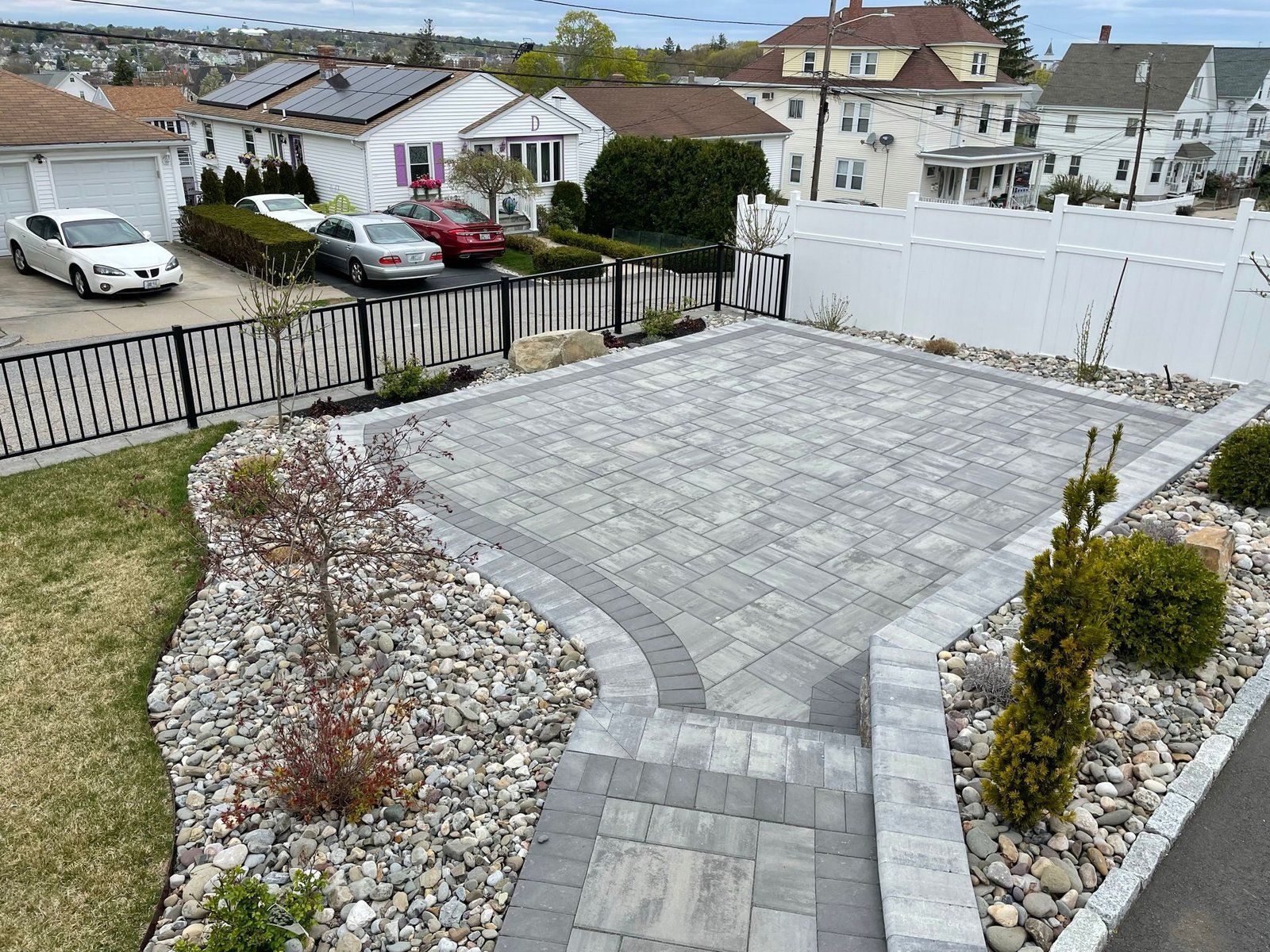 A stone patio and walkway featuring gray pavers with a darker border, surrounded by rocks and landscaping in a backyard.