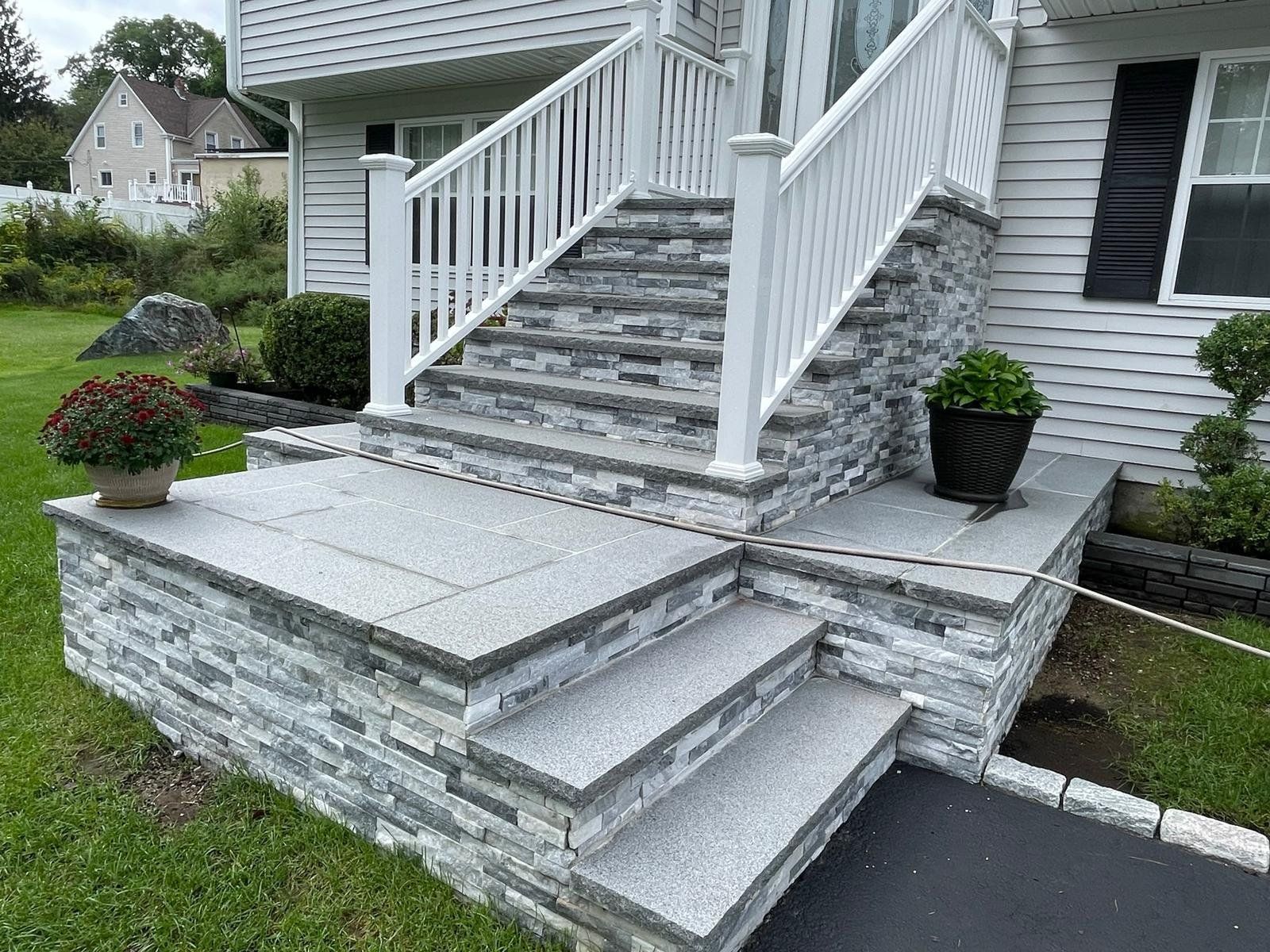 A stone front porch with granite steps, white railings, and a potted plant leading to the entrance of a grey house.