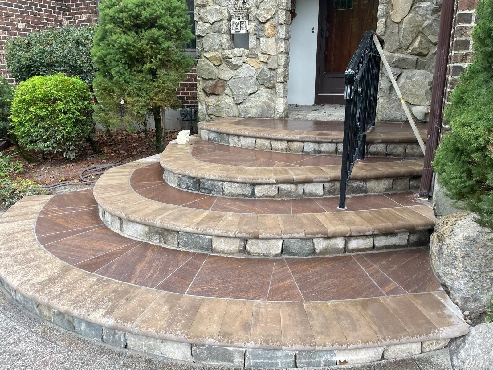 Curved stone steps leading to a building entrance with a decorative black metal railing and surrounding landscaping.