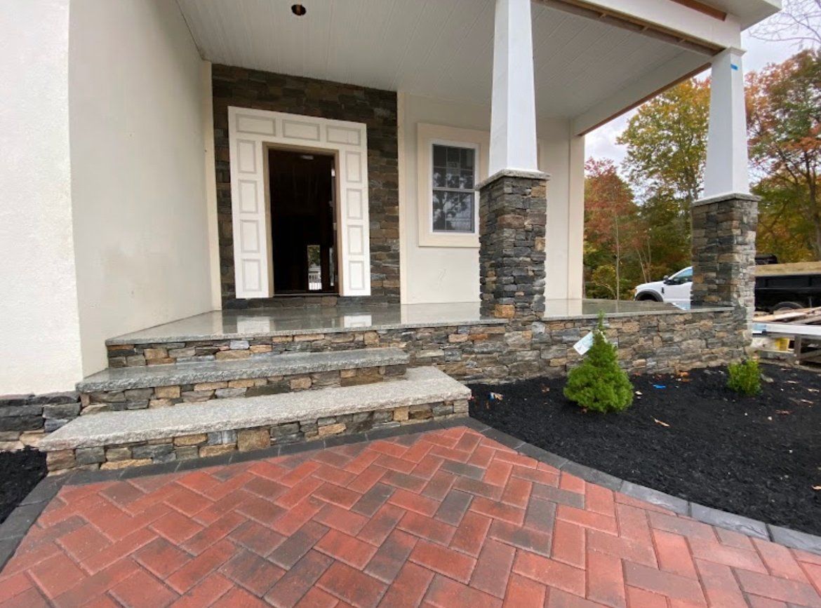A front porch with stone-veneer steps and columns, leading to a doorway, featuring a red brick herringbone walkway.