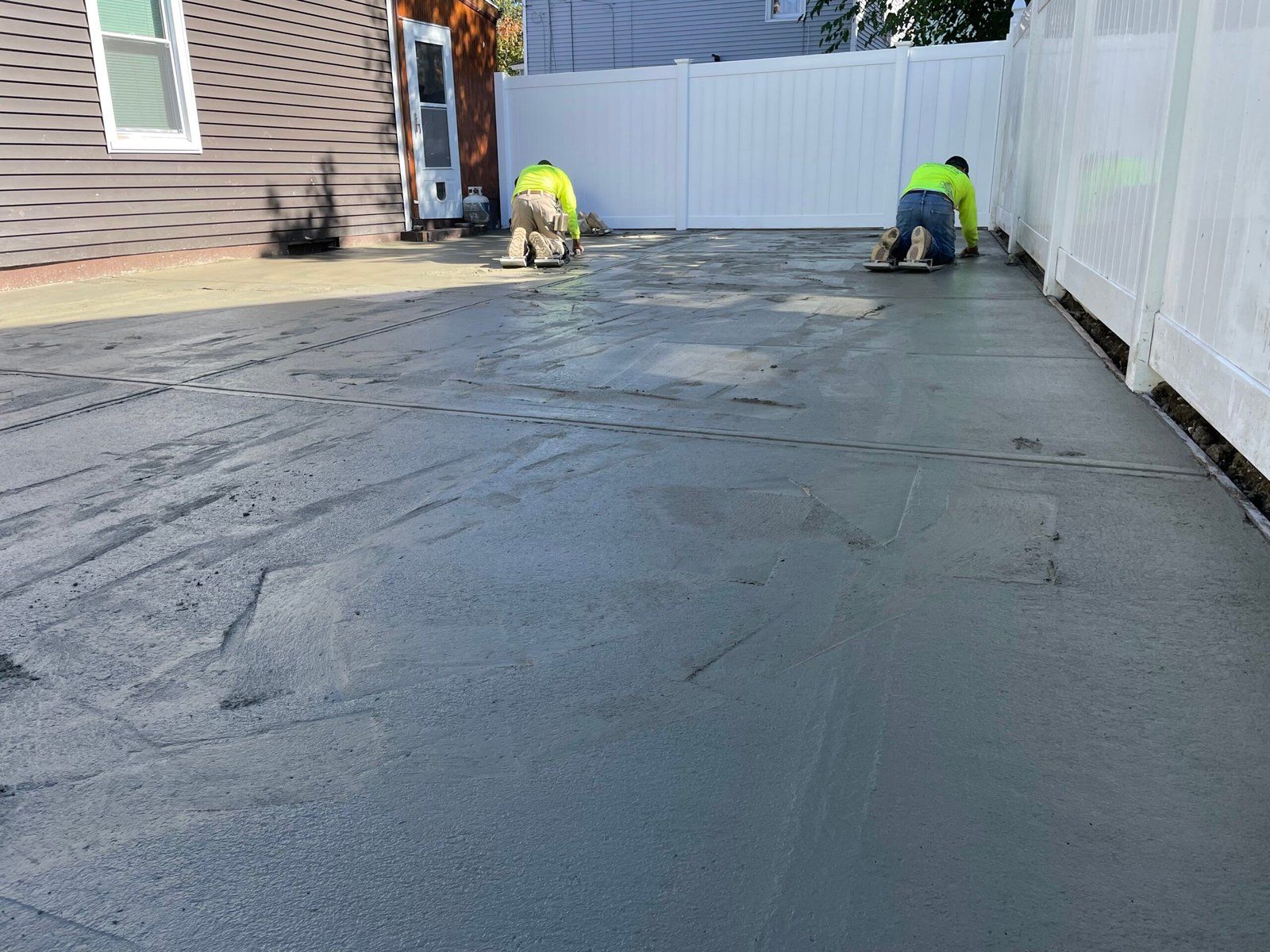 Two people in high-visibility neon shirts finish smoothing a fresh, wet concrete patio between a house and a white fence.