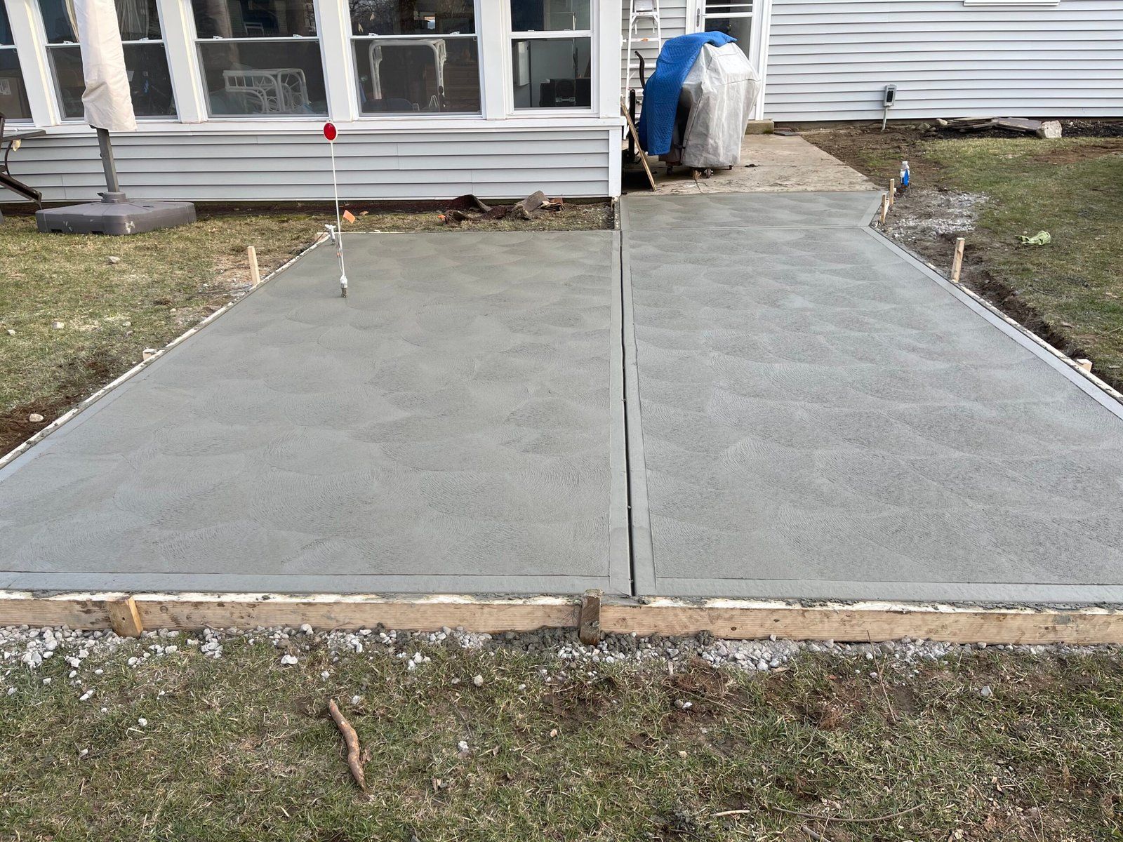A new rectangular concrete patio slab with wooden formwork surrounds, set against the side of a house.