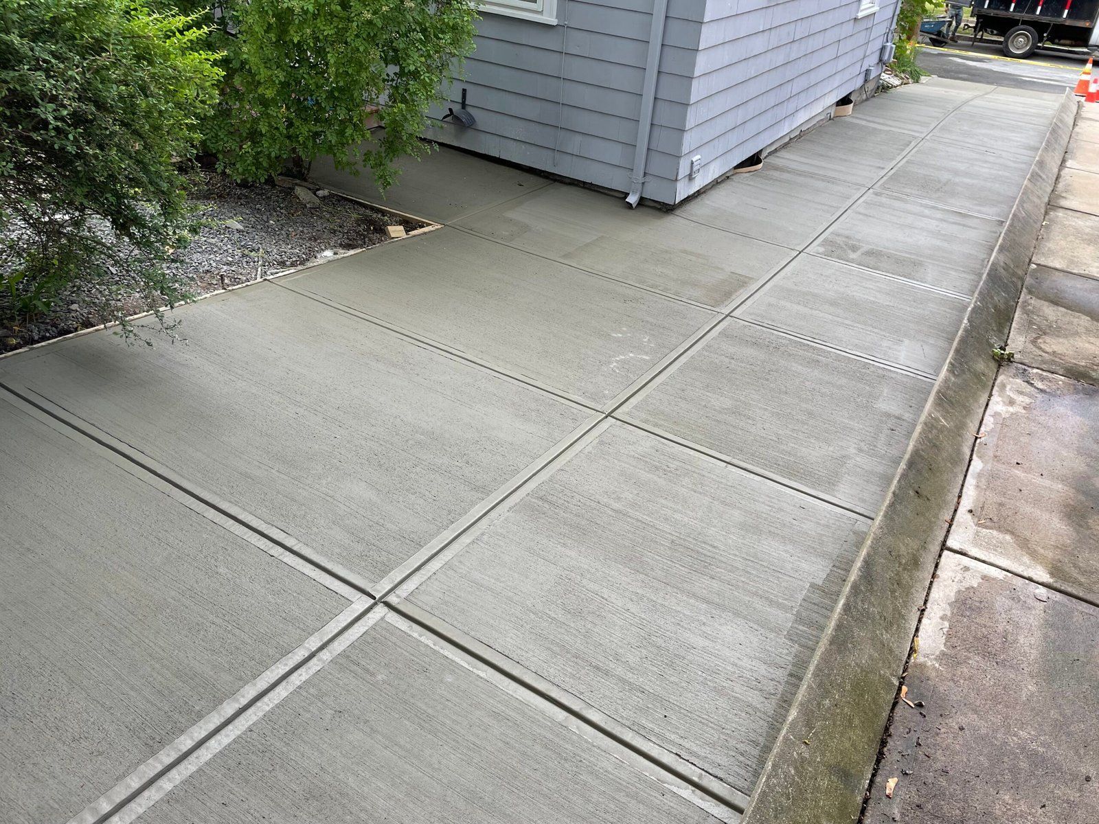 A newly poured, smooth concrete walkway running alongside the side of a house next to a patch of green bushes.