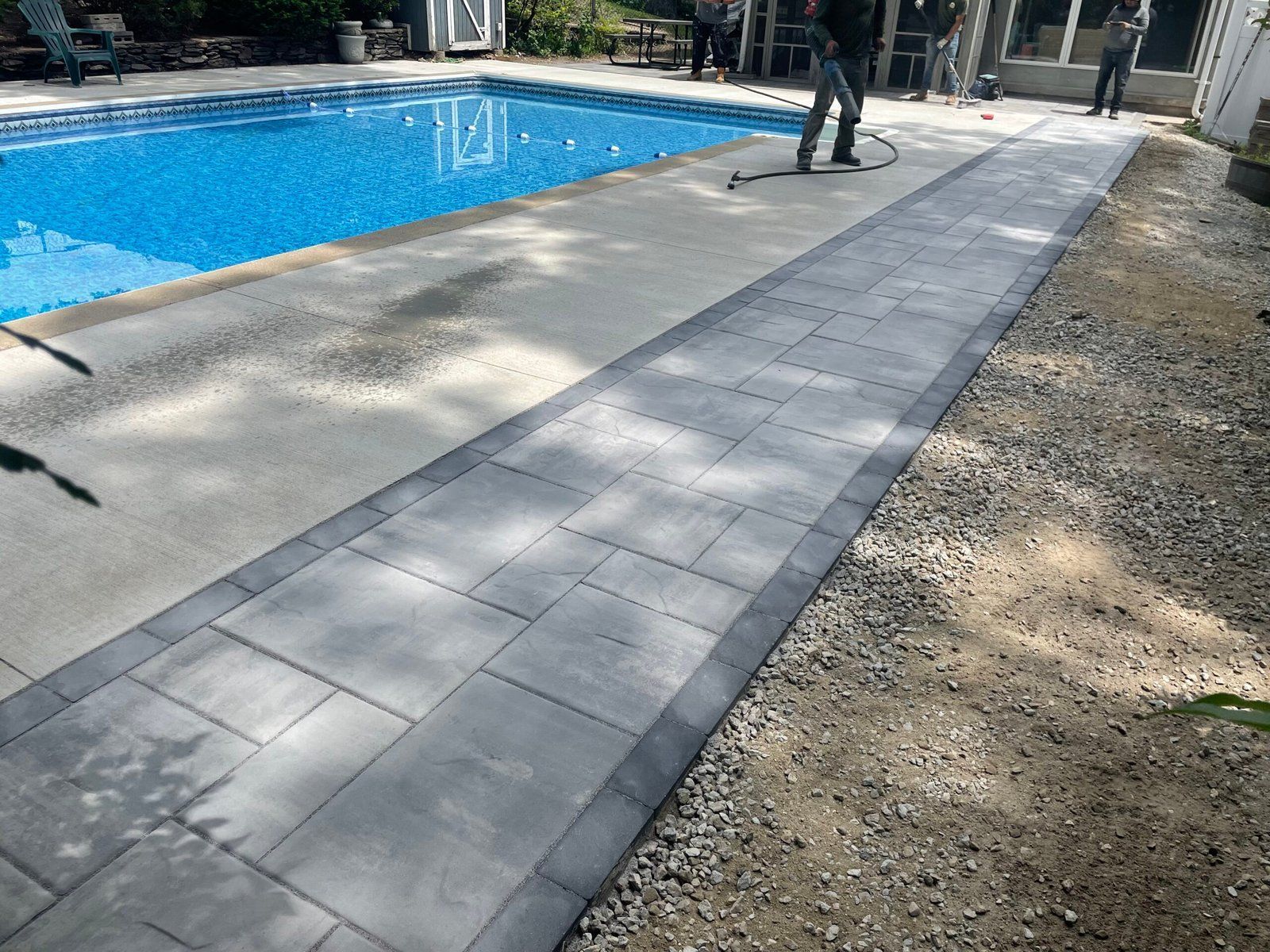 A paved walkway bordering a blue swimming pool, with gravel on the right and people standing in the background.