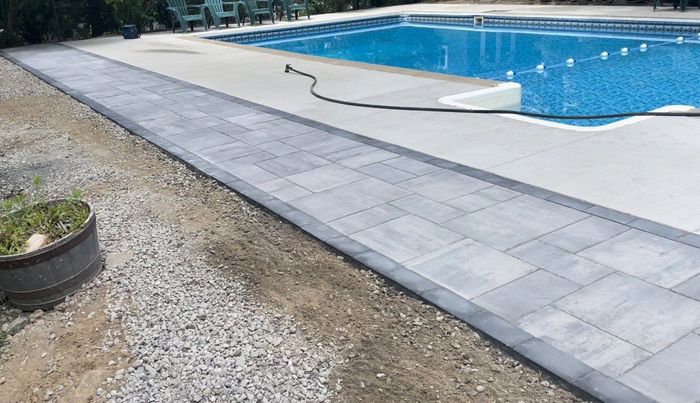 A paver walkway border sits between a concrete pool deck and a gravel yard.