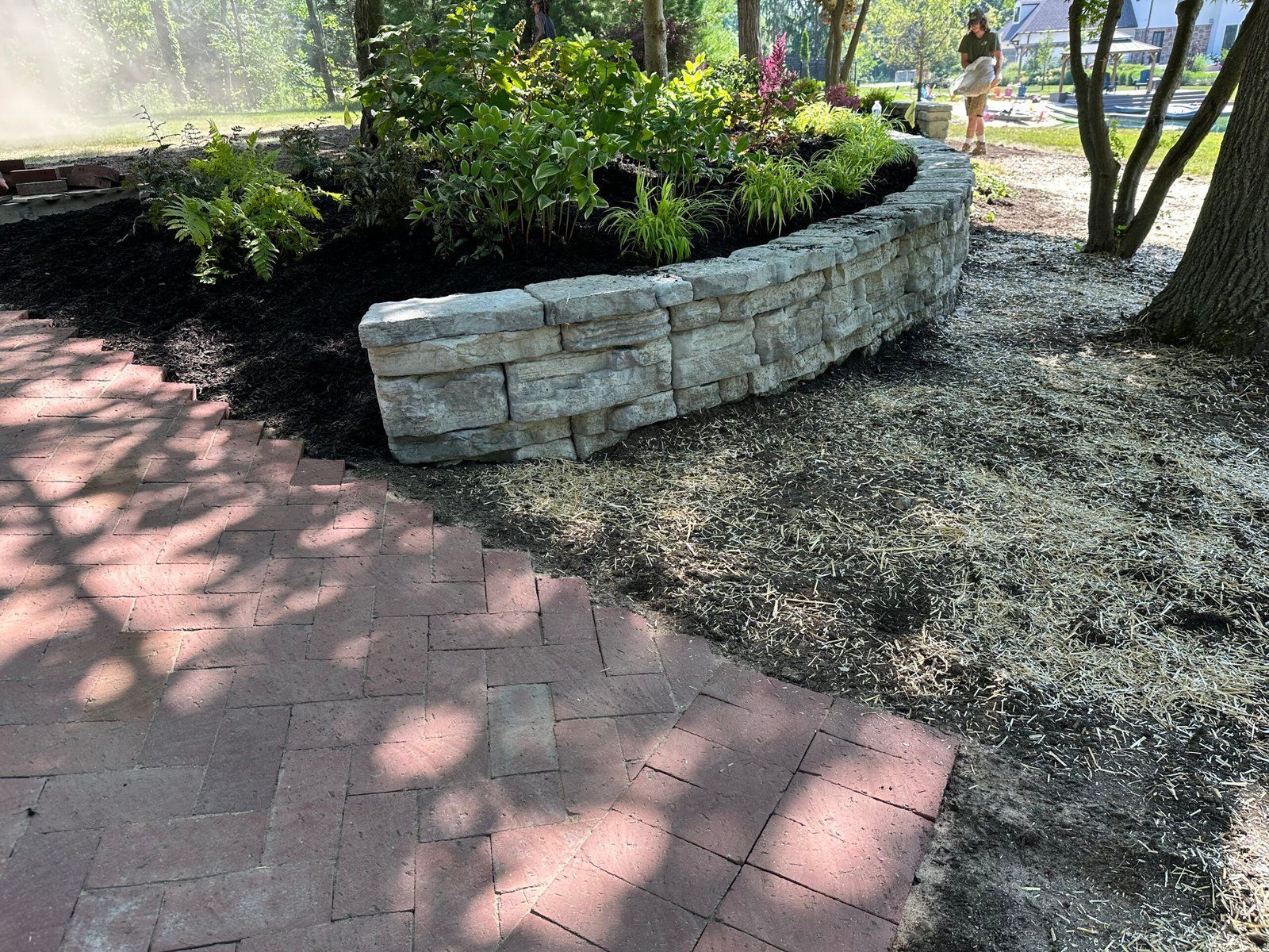 A curved stone retaining wall borders a garden bed next to a red brick patio in a sunny yard.