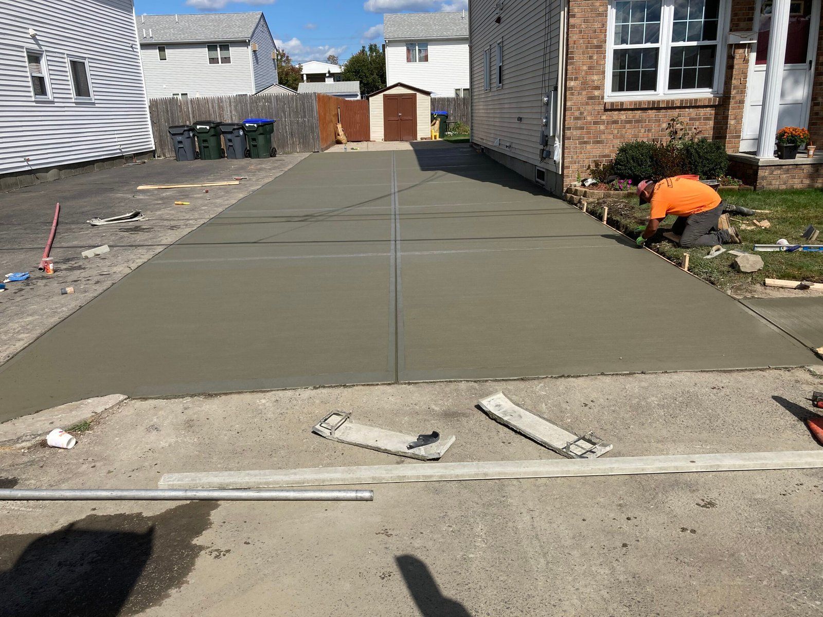 A worker in an orange shirt finishes a freshly poured concrete driveway beside a residential house.