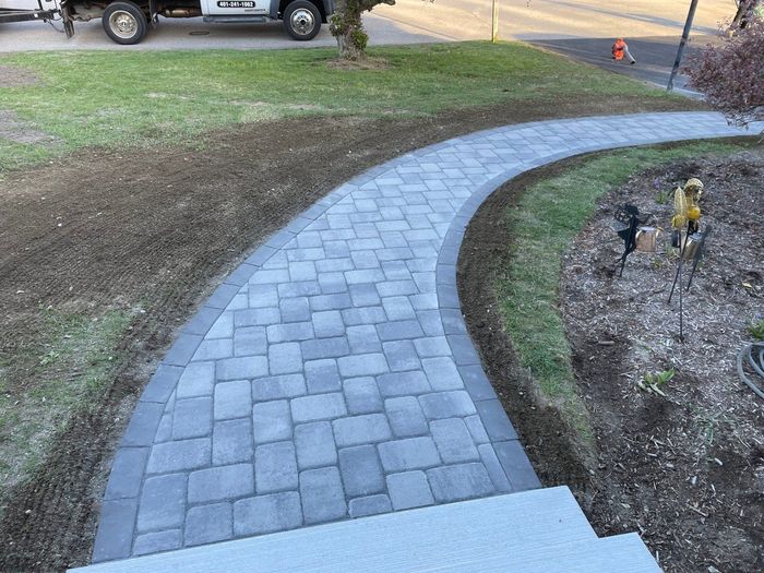 A curved walkway made of grey rectangular pavers with a dark border, bordered by grass and a mulched garden bed.