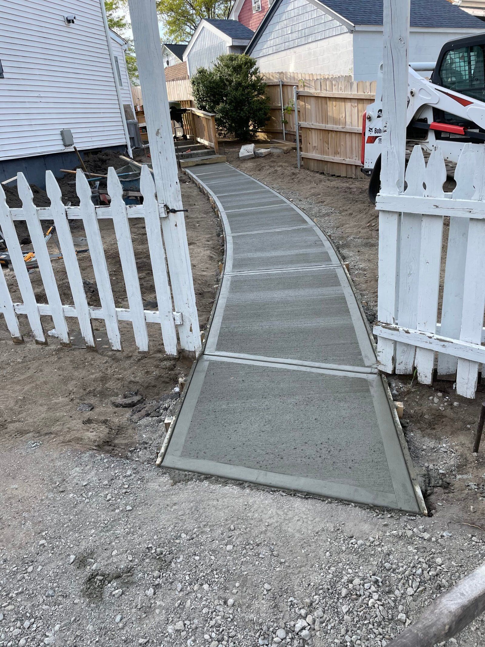 A newly poured concrete walkway curves between two sections of a white picket fence toward a house.