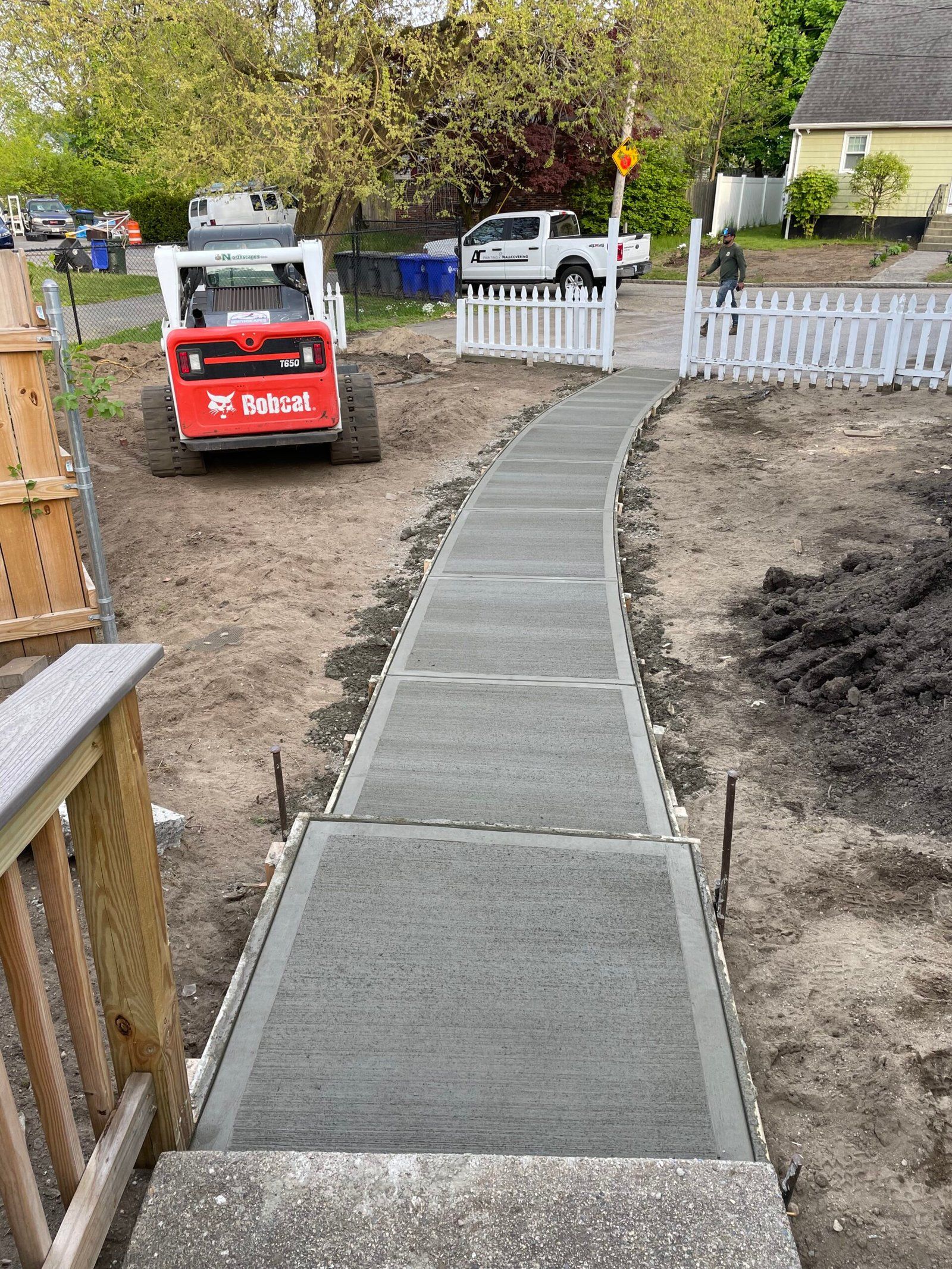 A freshly poured concrete sidewalk leads from a wooden porch toward a white picket fence, with a red skid steer nearby.