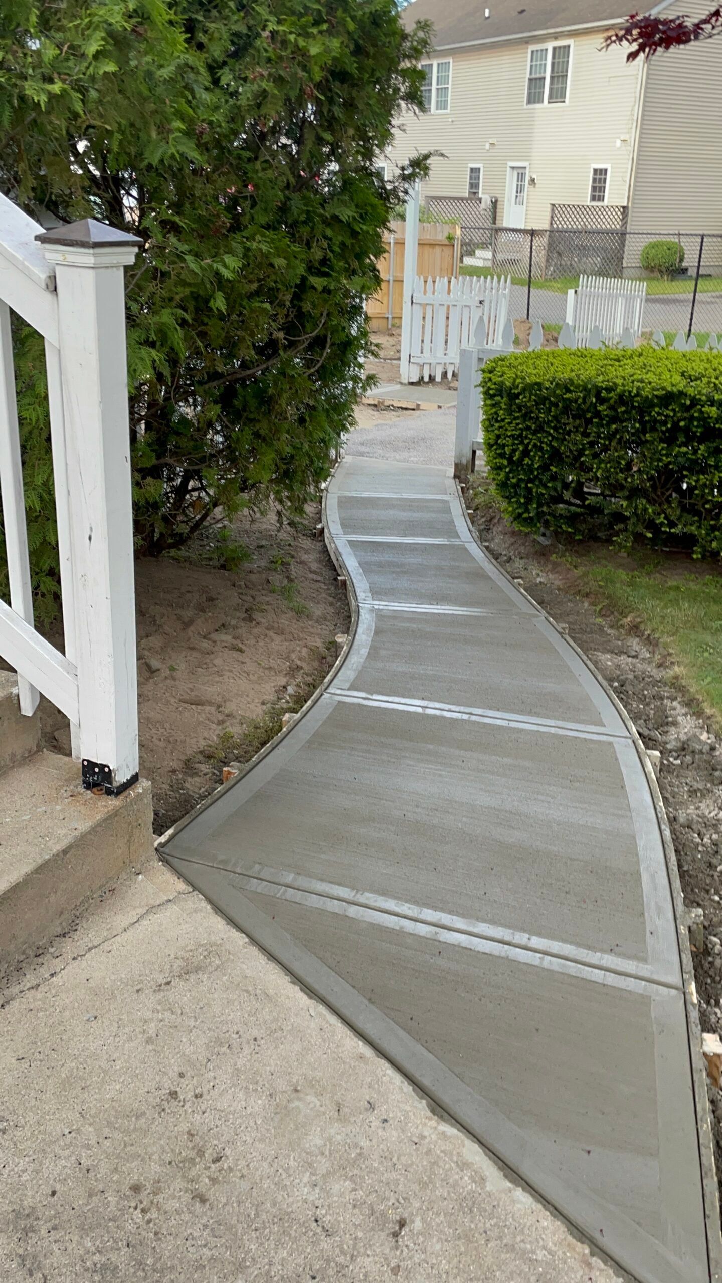 A newly poured, curved concrete walkway leading from steps toward a white fence in a residential yard.
