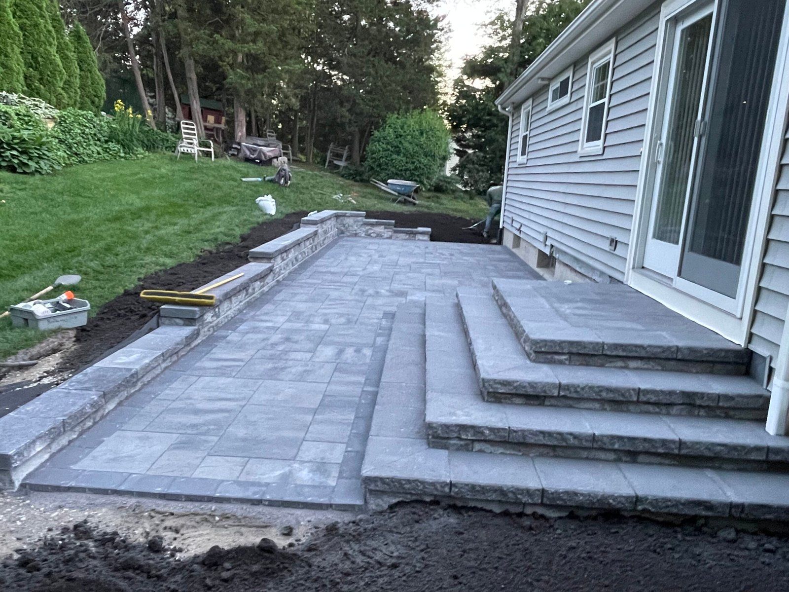New gray stone patio and tiered steps leading to a residential sliding door, with a lawn and trees in the background.