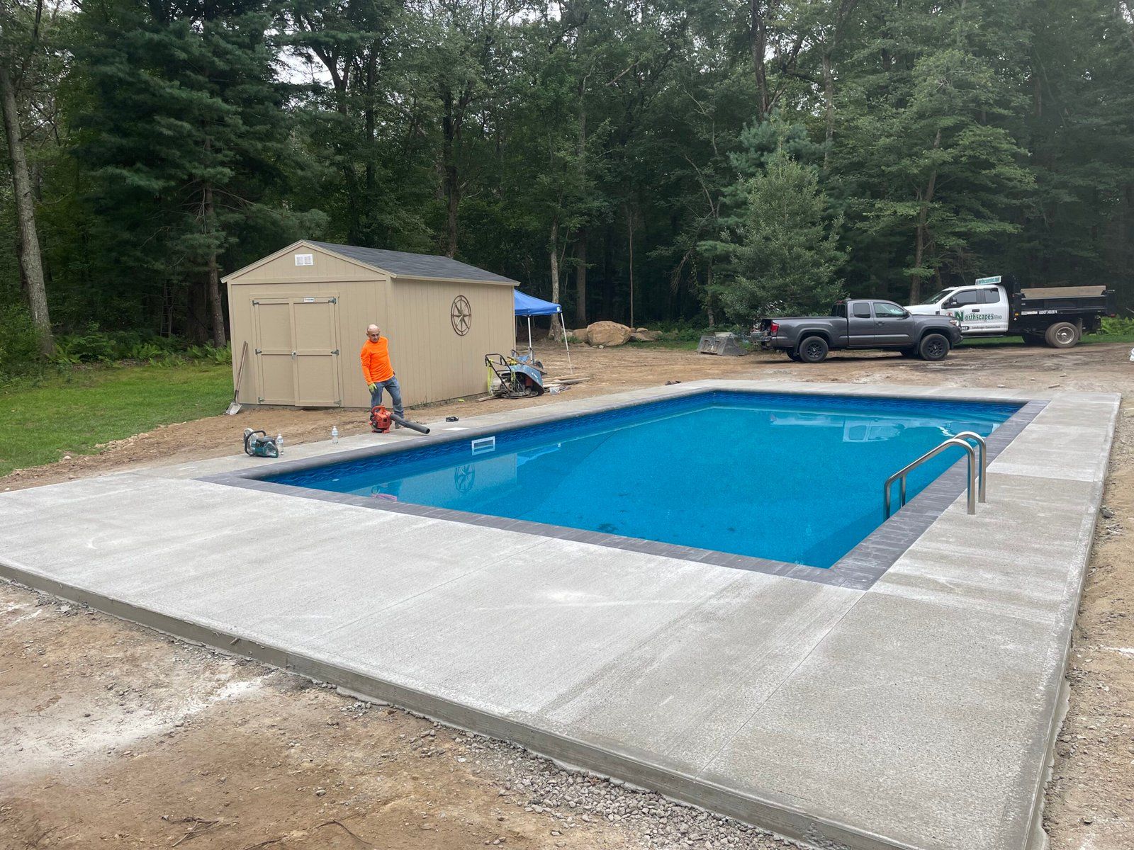 A rectangular inground pool with a concrete deck under construction, with a storage shed and trucks in the background.