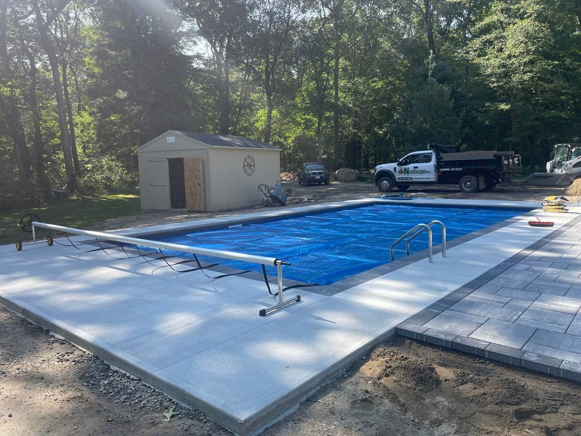 A rectangular backyard swimming pool with a blue solar cover, surrounded by new light-colored concrete and stone paving.