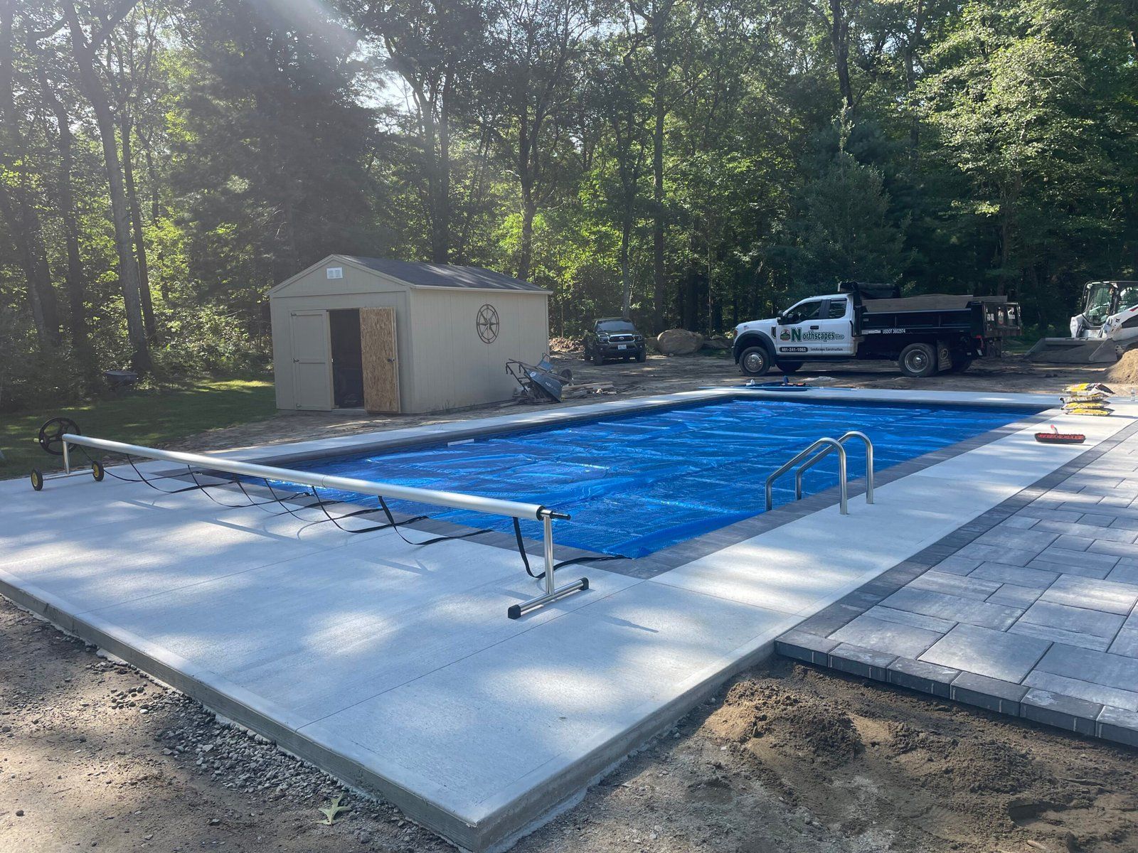 A rectangular backyard swimming pool covered with a blue tarp, surrounded by a light-colored concrete deck and pavers.