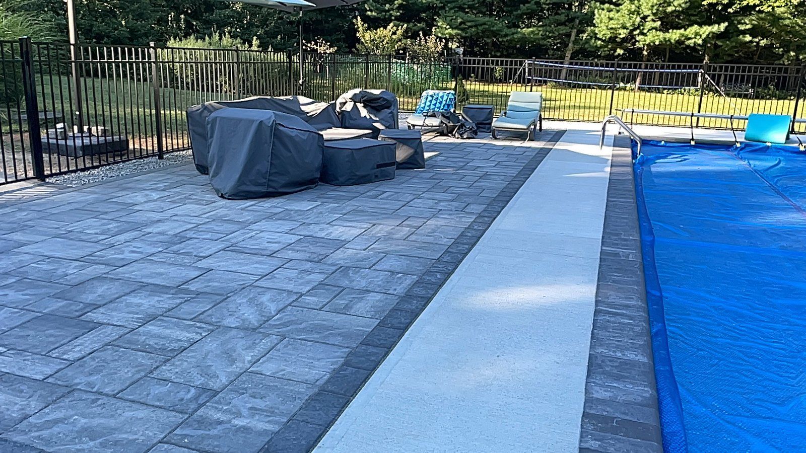 A grey paver patio with covered outdoor furniture, a white walkway, and a swimming pool covered with a blue solar blanket.