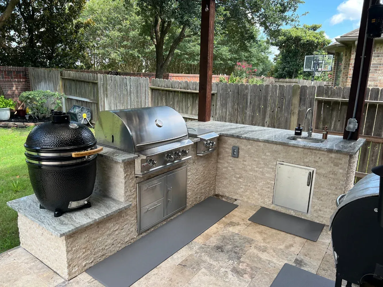 Outdoor kitchen with a stone-clad island, featuring a stainless steel gas grill, a black ceramic grill, and a small sink.