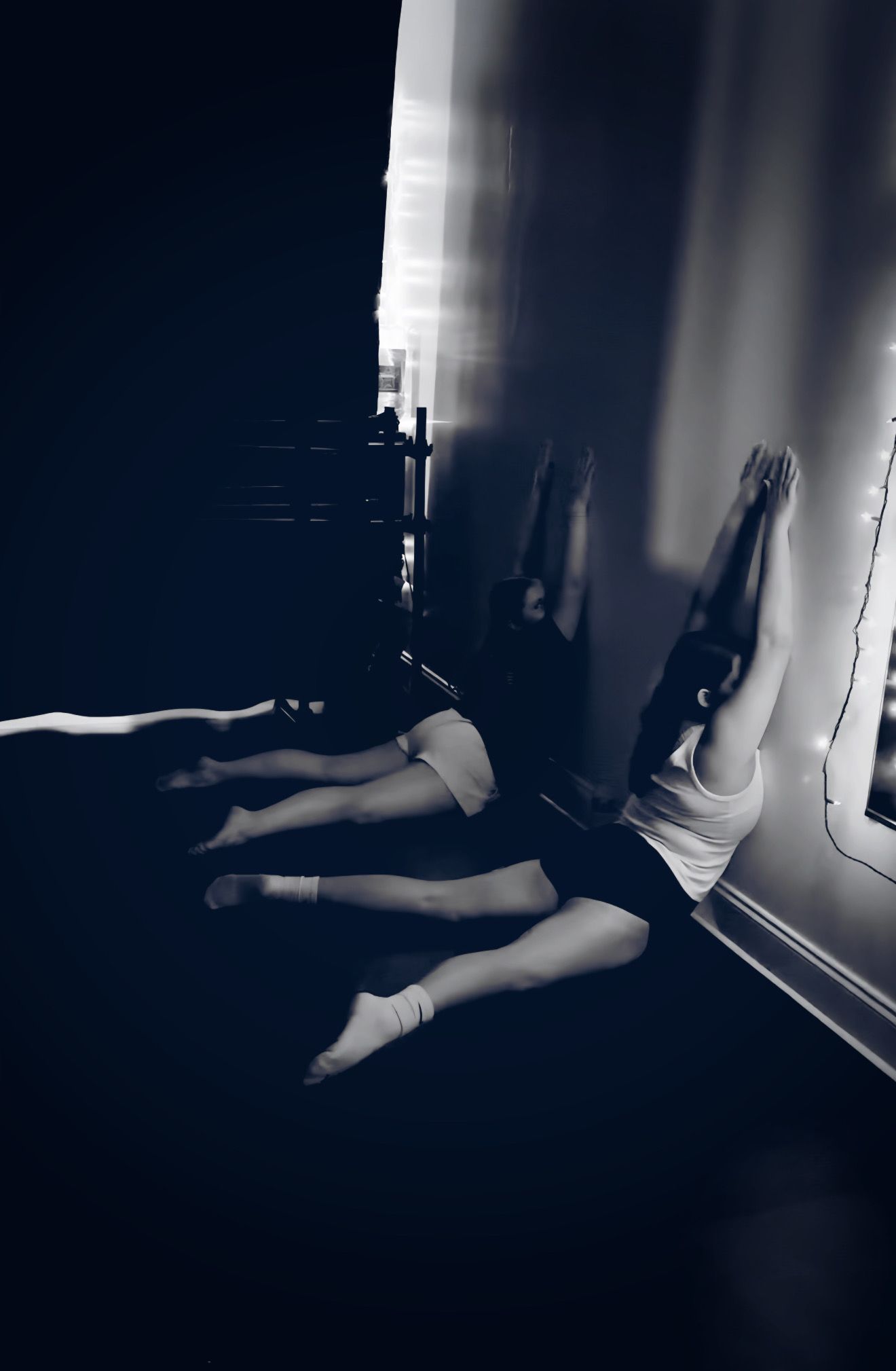 Two figures, legs extended, against a wall. They appear to be stretching on a dark floor.