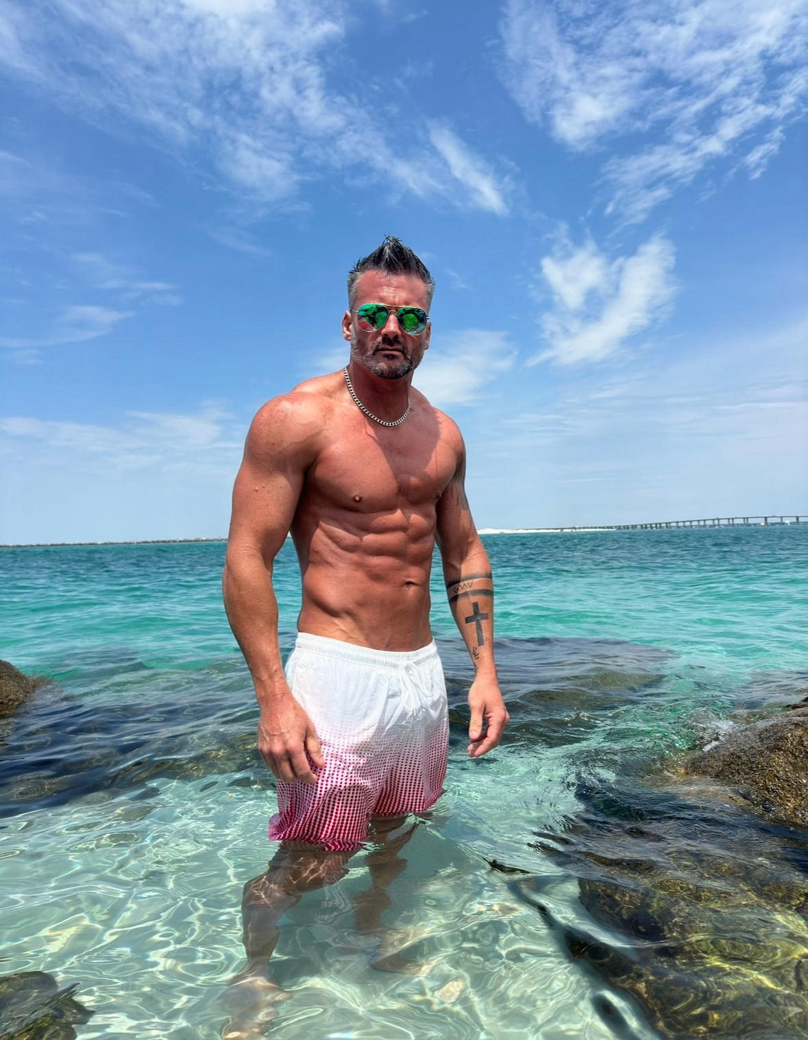 A shirtless man is standing in the ocean wearing sunglasses.