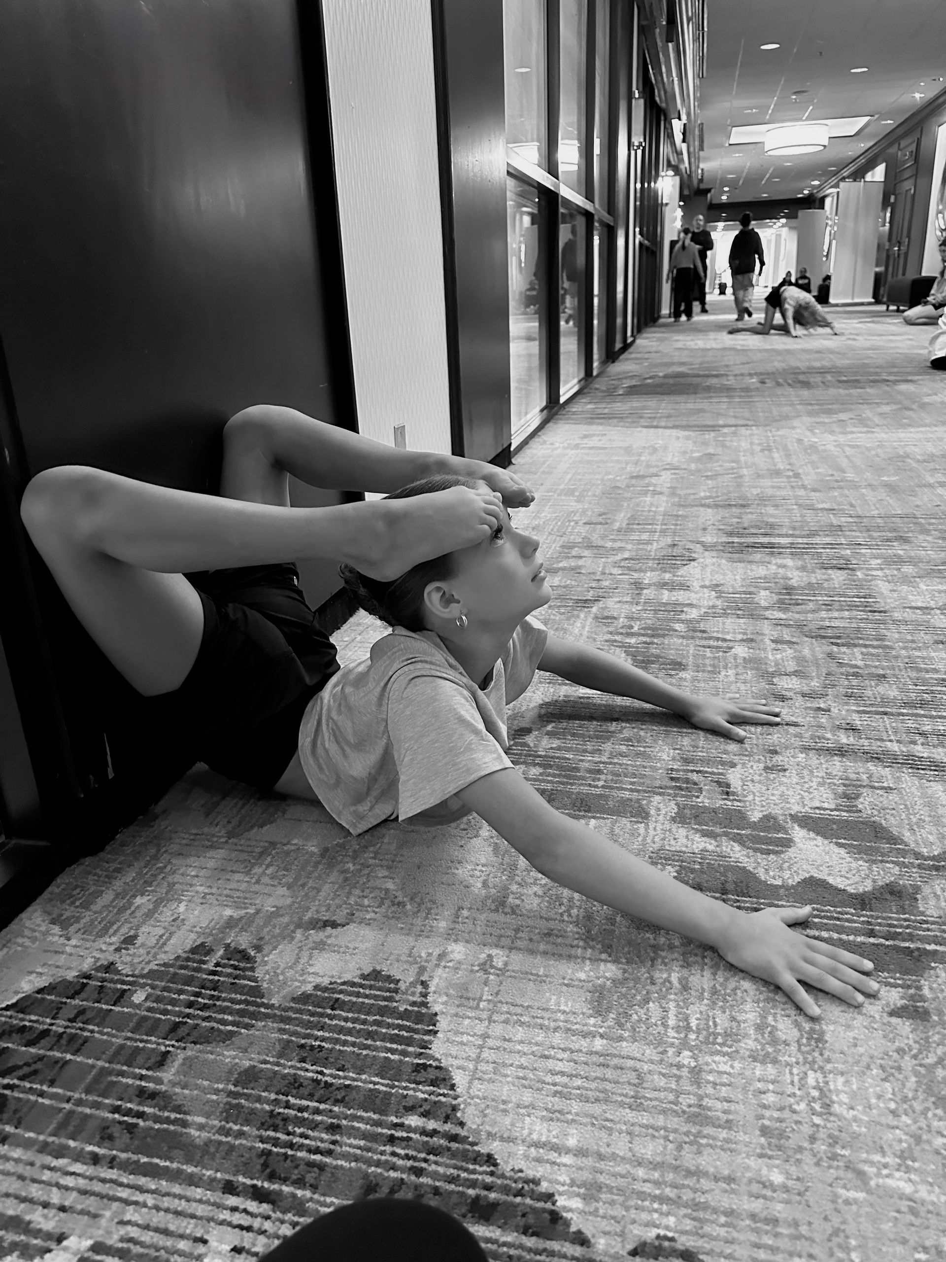 Person in a backbend on a carpeted floor, arms outstretched, legs bent over head. Long hallway in background.