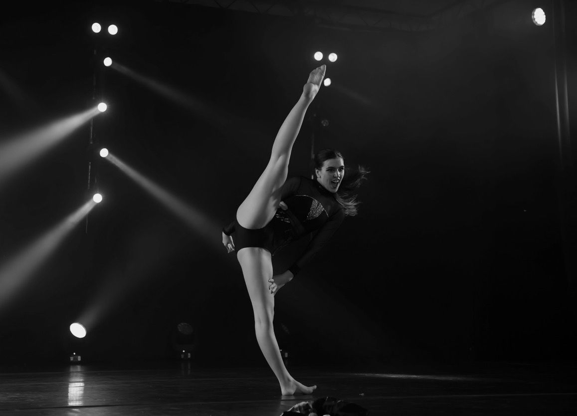 Dancer in a black leotard performs a high leg extension on stage, illuminated by spotlights.