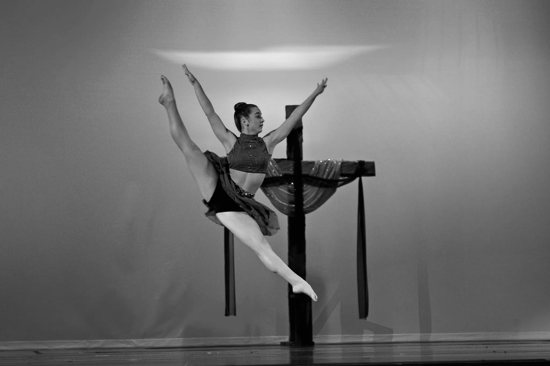 Dancer leaps with arms outstretched, leg extended. Backdrop: cross with draped fabric. Black and white photo.