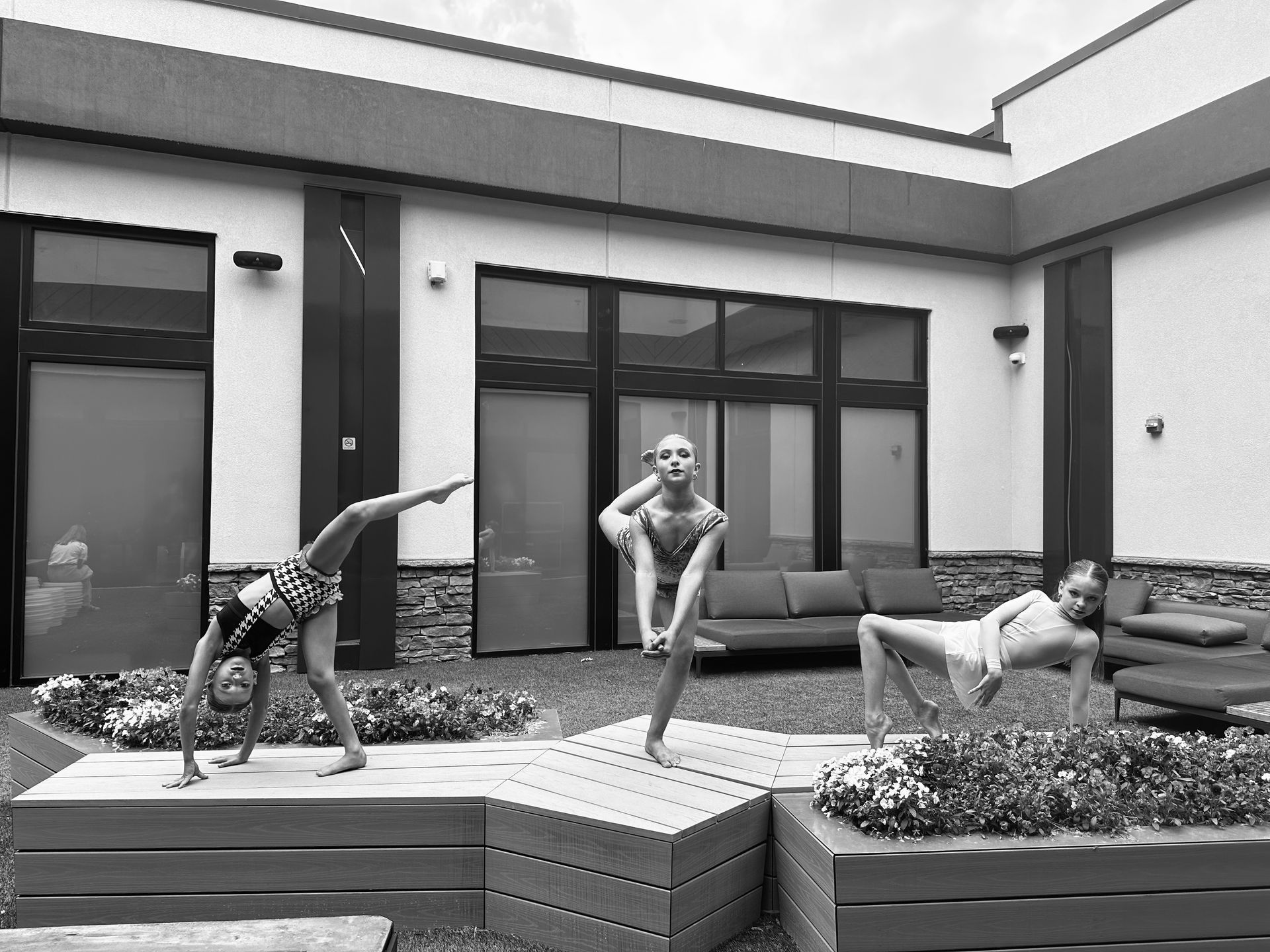Three girls doing acrobatic moves on a patio with a building backdrop and plants.