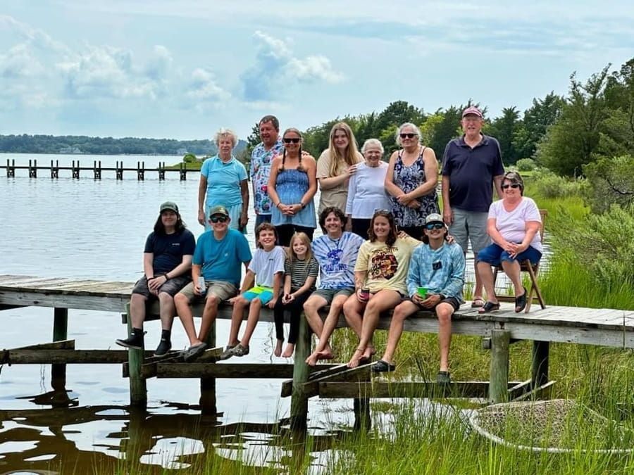 First Landing team posing on a wooden dock by a lake, with water, trees, and a blue sky in the background.