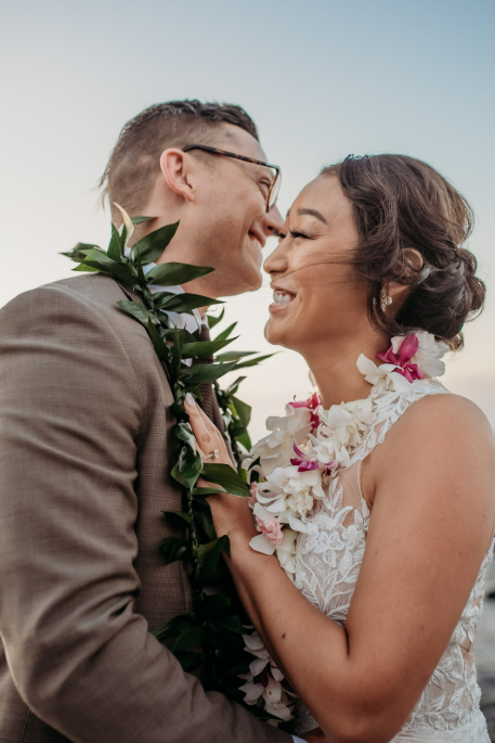 Couple wearing leis on beach at sunset. Woman smiles, holds flowers; man in hat smiles.