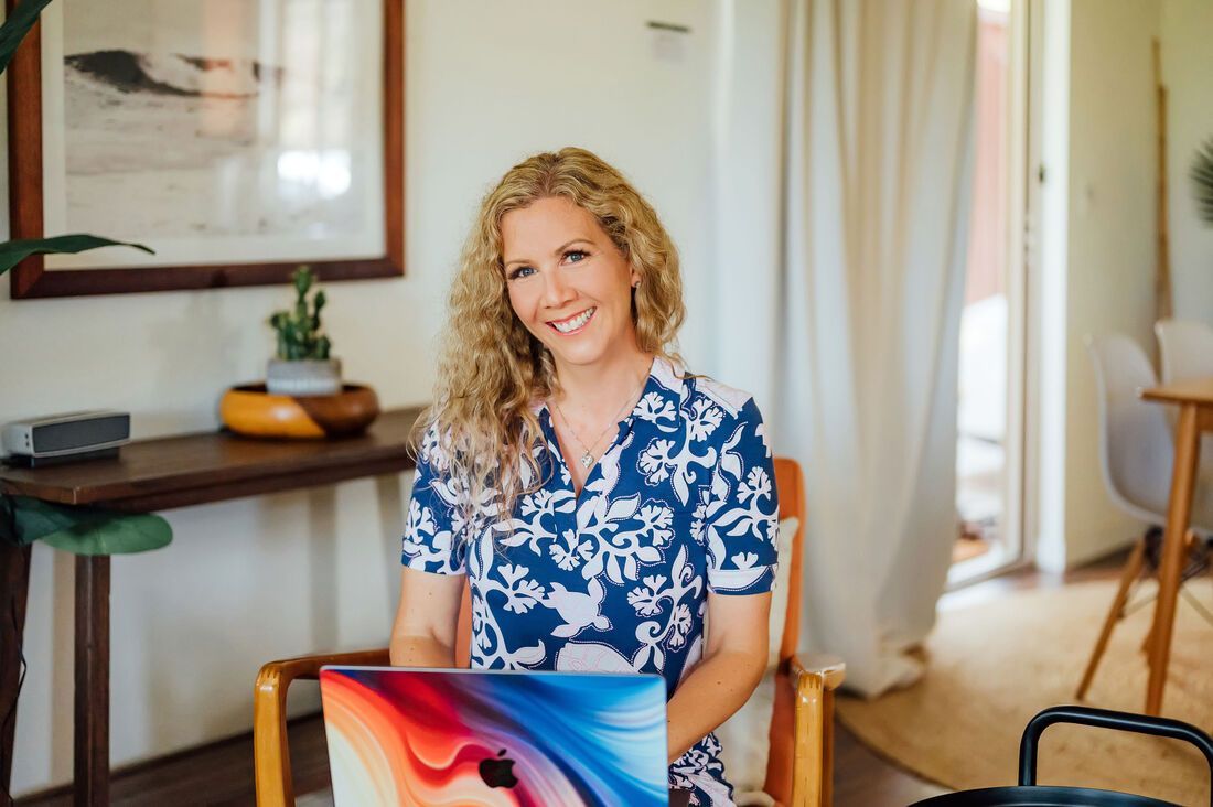 Woman with curly blonde hair smiles, seated with laptop in a room with wood table, plants, and natural light.