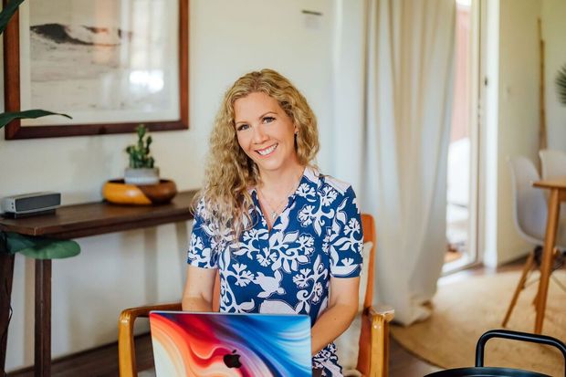 Woman with curly blonde hair smiles, seated with laptop in sunlit room.