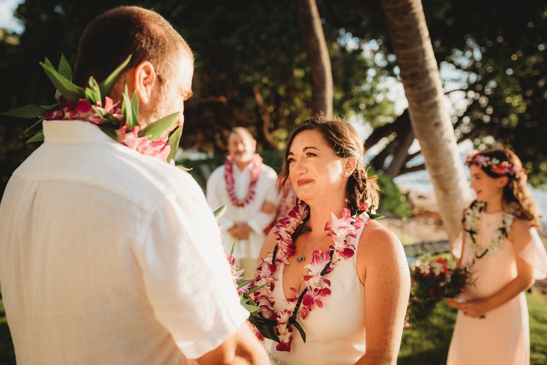 Couple exchanging vows during a tropical wedding ceremony, wearing leis. Attendees in the background.