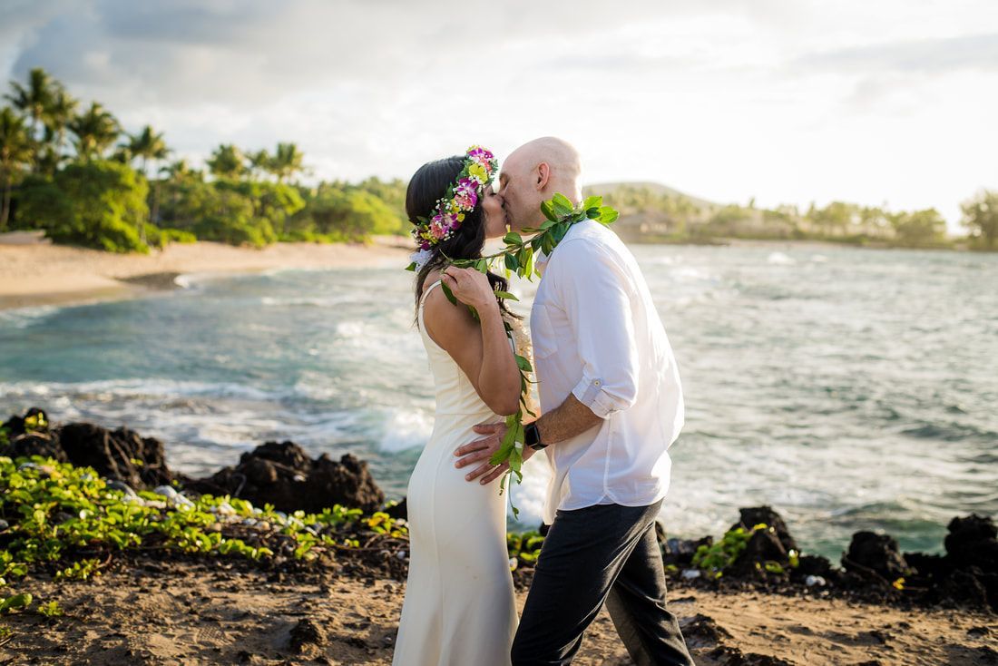 Couple kissing on a beach. Bride wears a floral crown, groom has a lei. Ocean and palm trees in background.