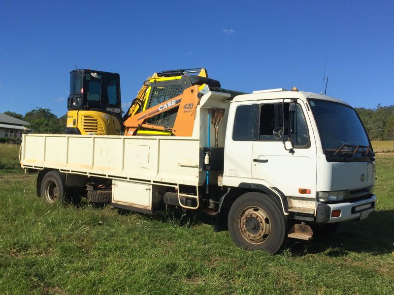 A white truck with a yellow excavator in the back is parked in a grassy field.