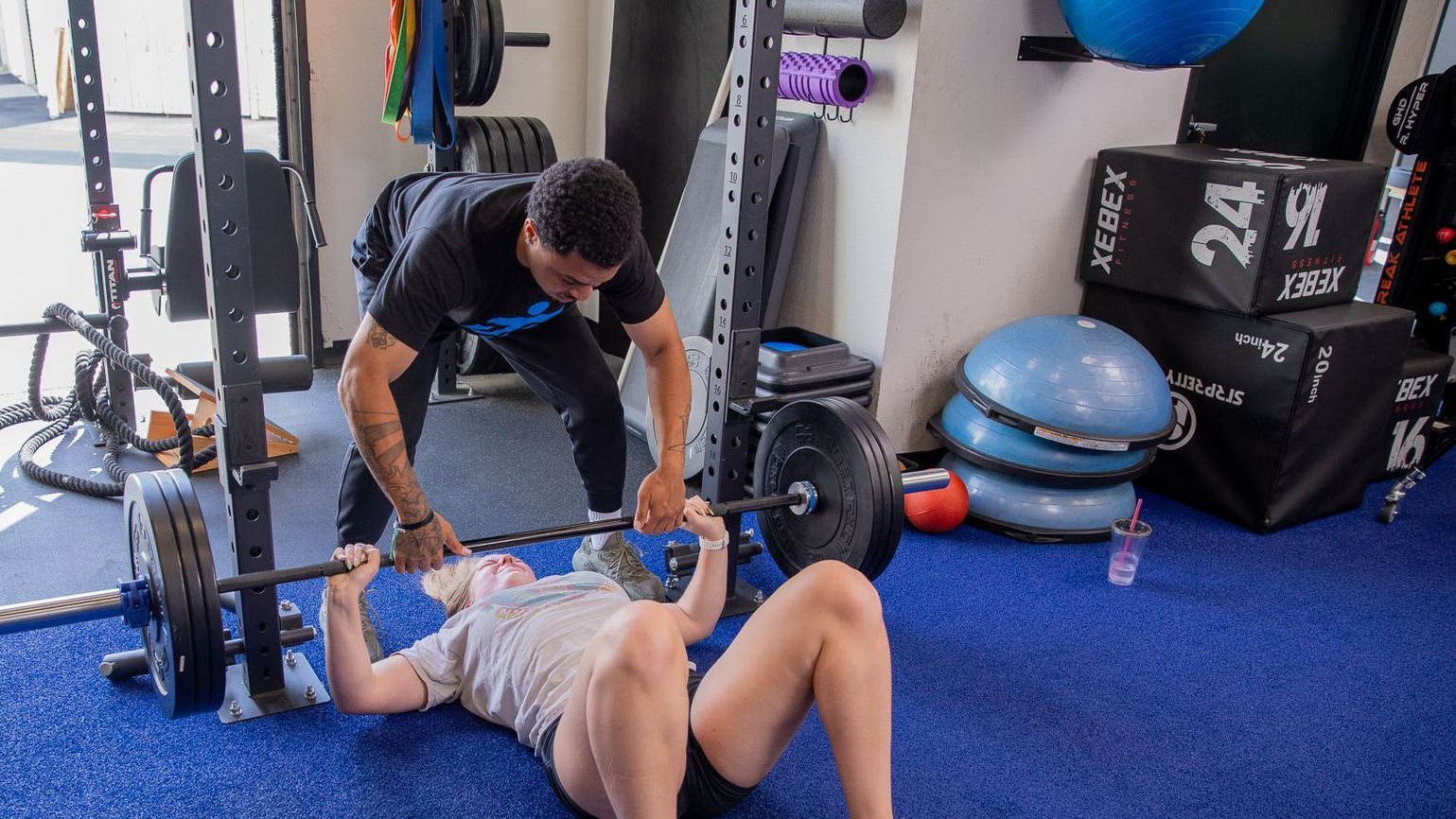 A man is helping a woman lift a barbell in a gym.