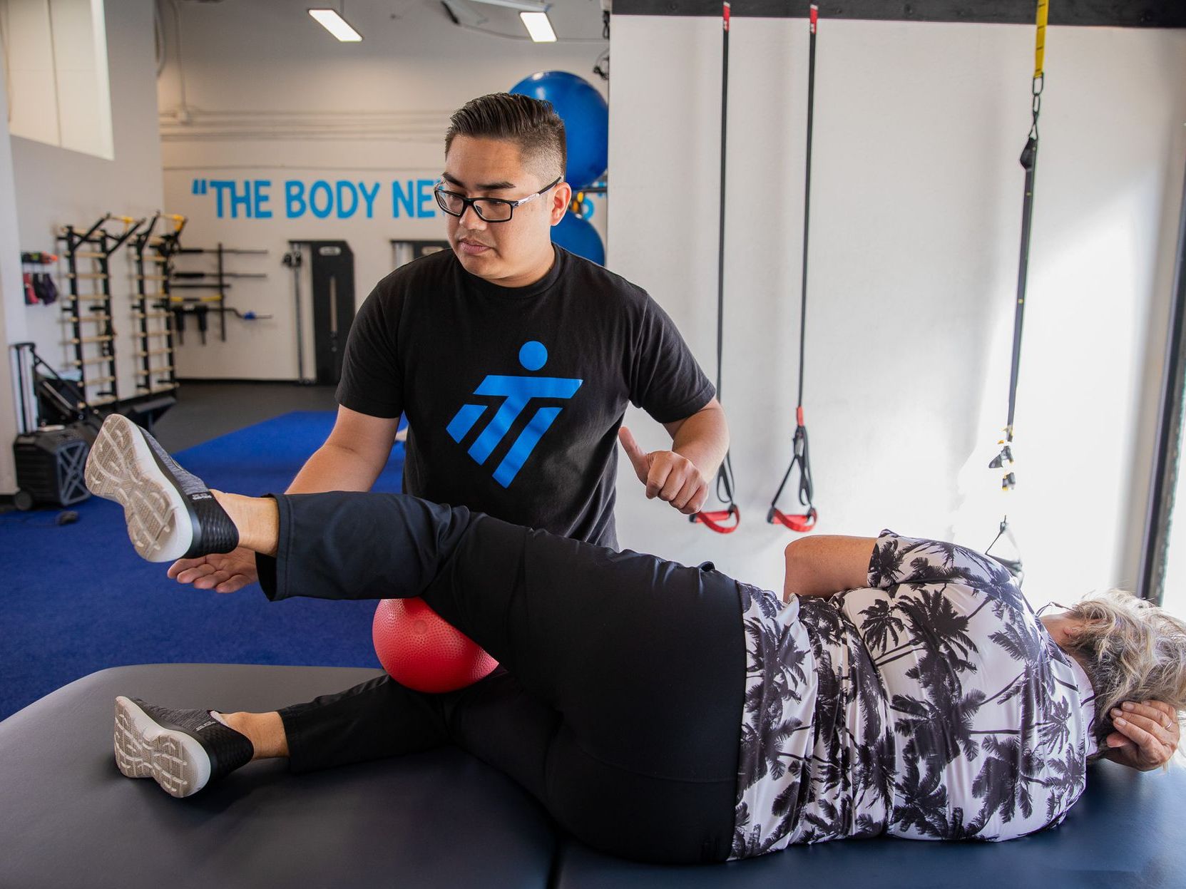 A man is helping a woman stretch her legs in a gym.