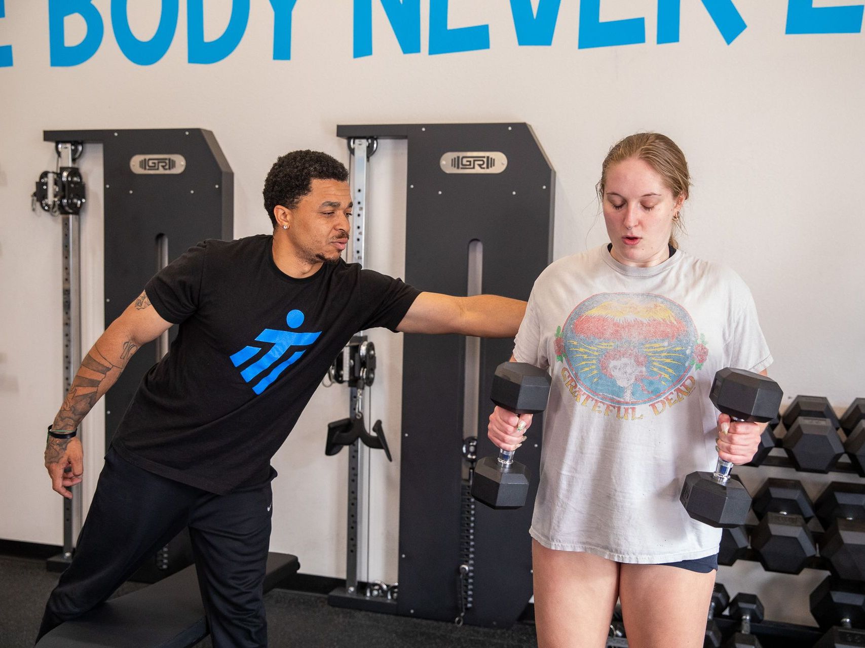 A man is helping a woman lift dumbbells in a gym.