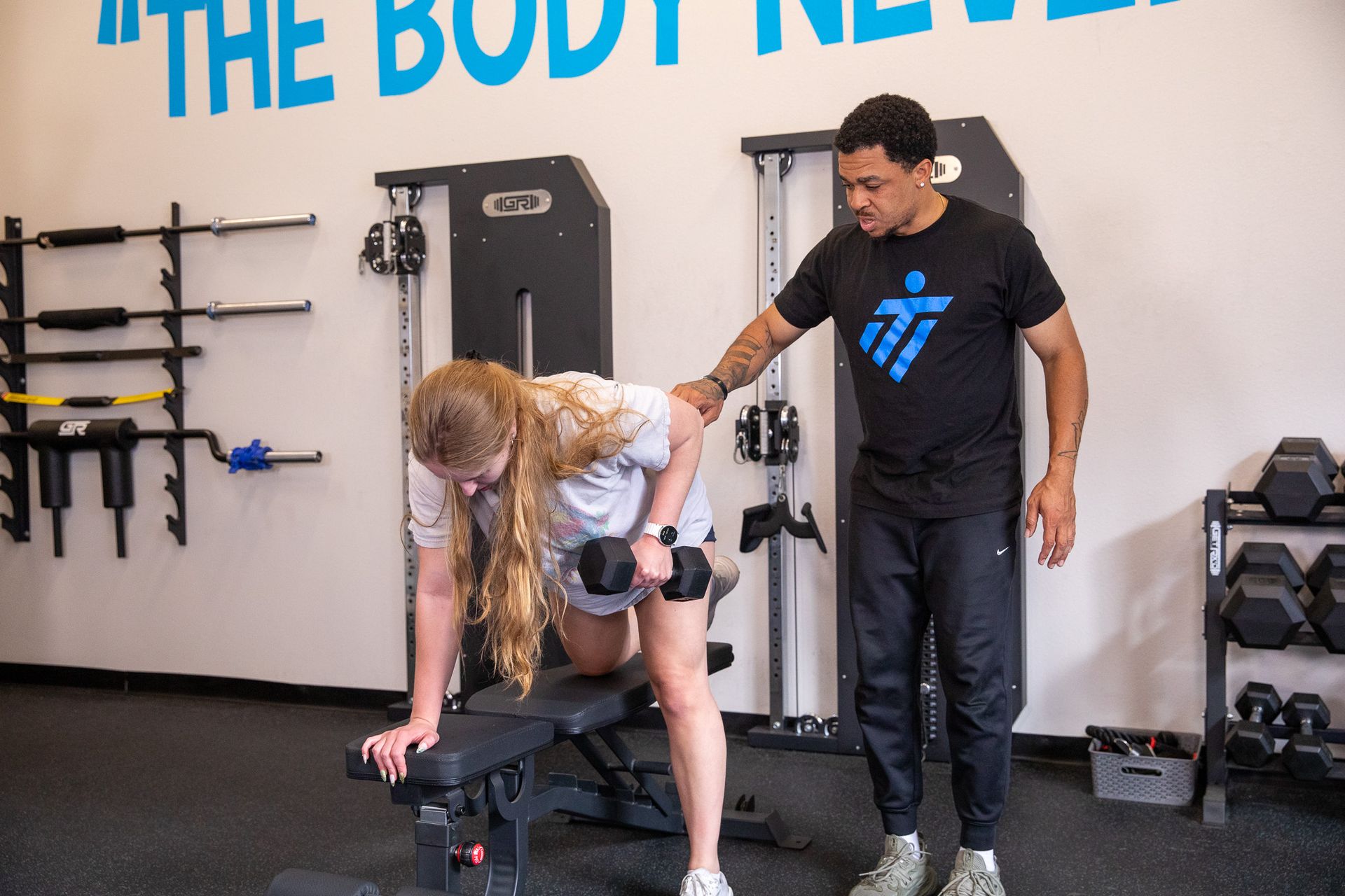 Woman doing dumbbell row, guided by trainer. In gym.