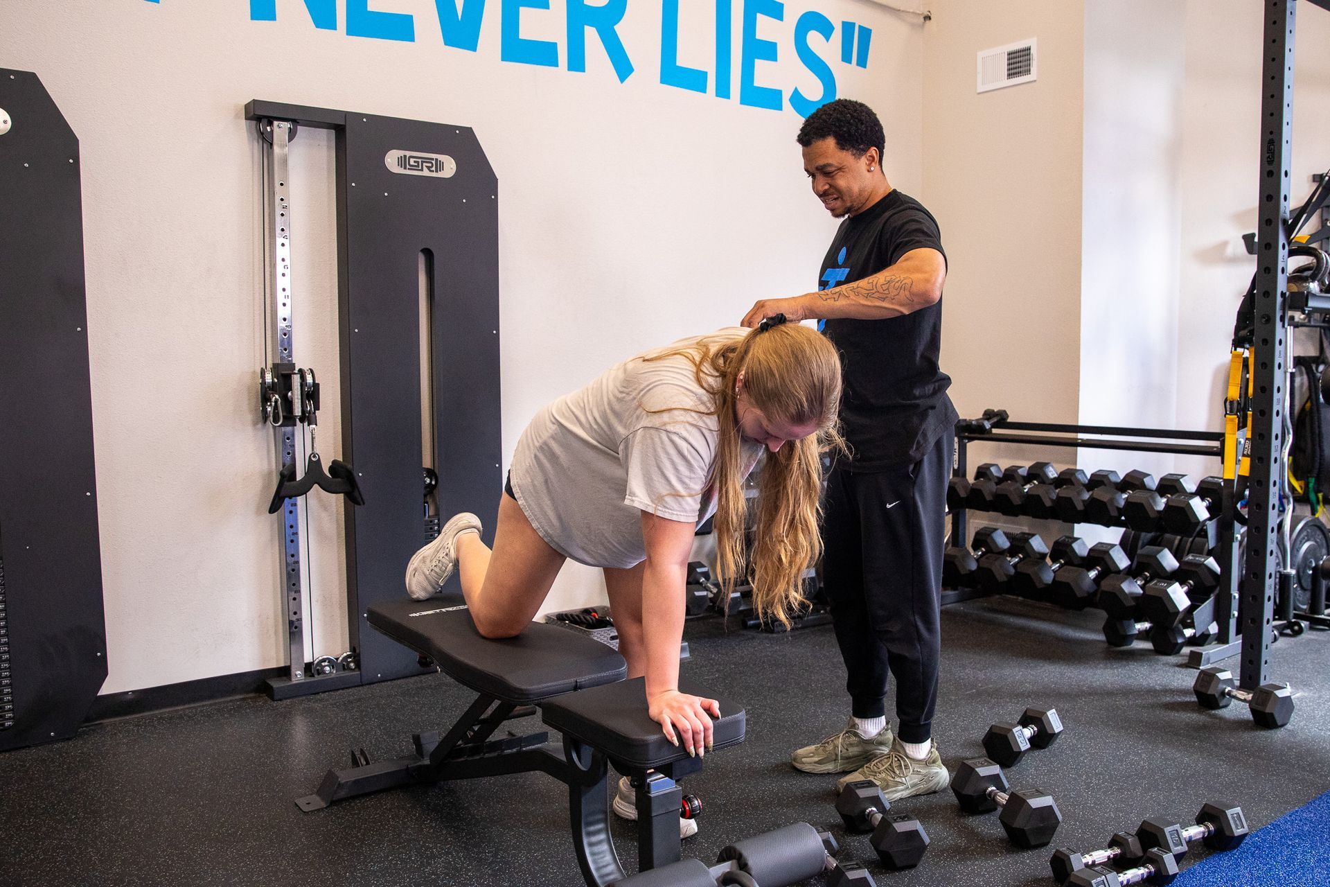A man is helping a woman do exercises on a bench in a gym.