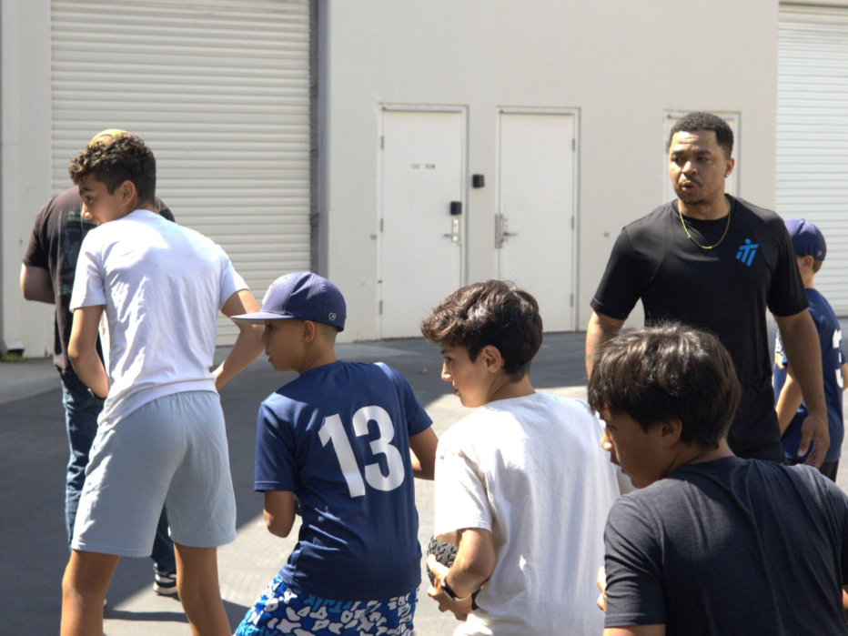 Group of boys playing outside, coached by a man. Boys wear t-shirts and hats; one wears a jersey with the number 13.