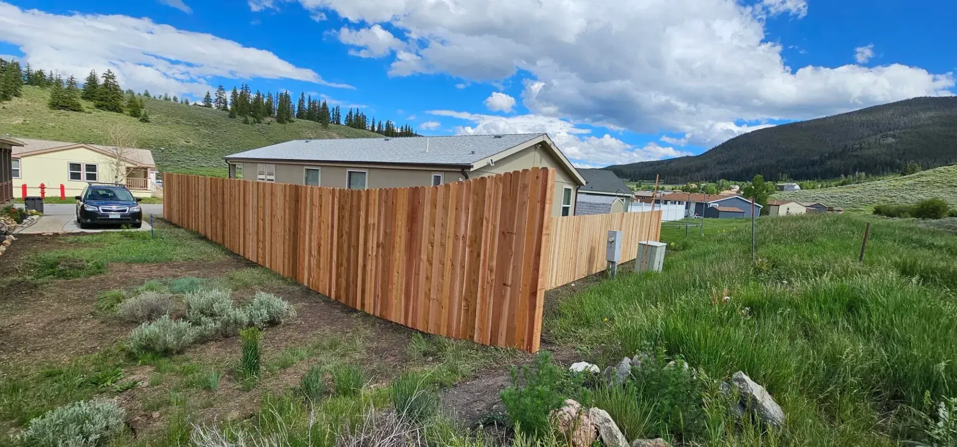 Wooden fenced yard beside a small building under a blue sky with clouds, mountains in the distance