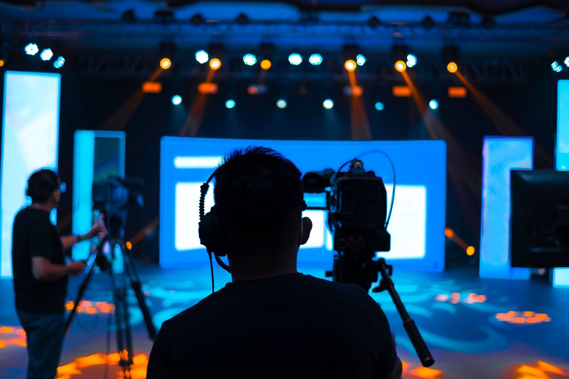 Silhouette of a person with headphones and camera filming a stage with blue backdrop and lights.