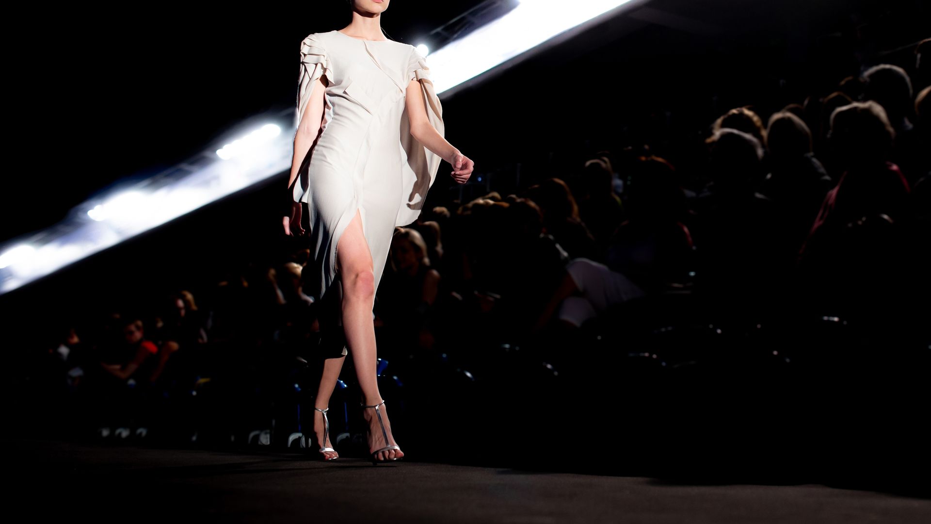Model in white dress walks the runway at a fashion show, lit by overhead spotlights, crowd in background.