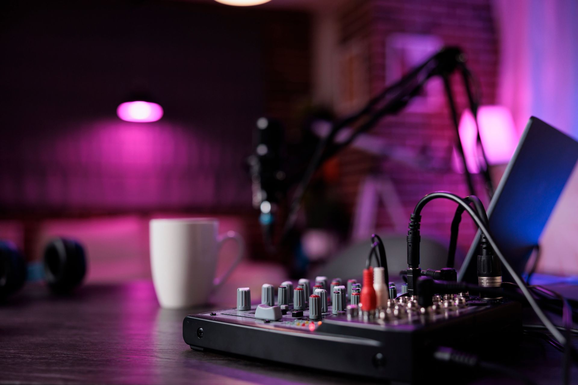 Audio mixing board with cables, microphone, laptop, mug, and headphones on a desk, bathed in purple and pink light.