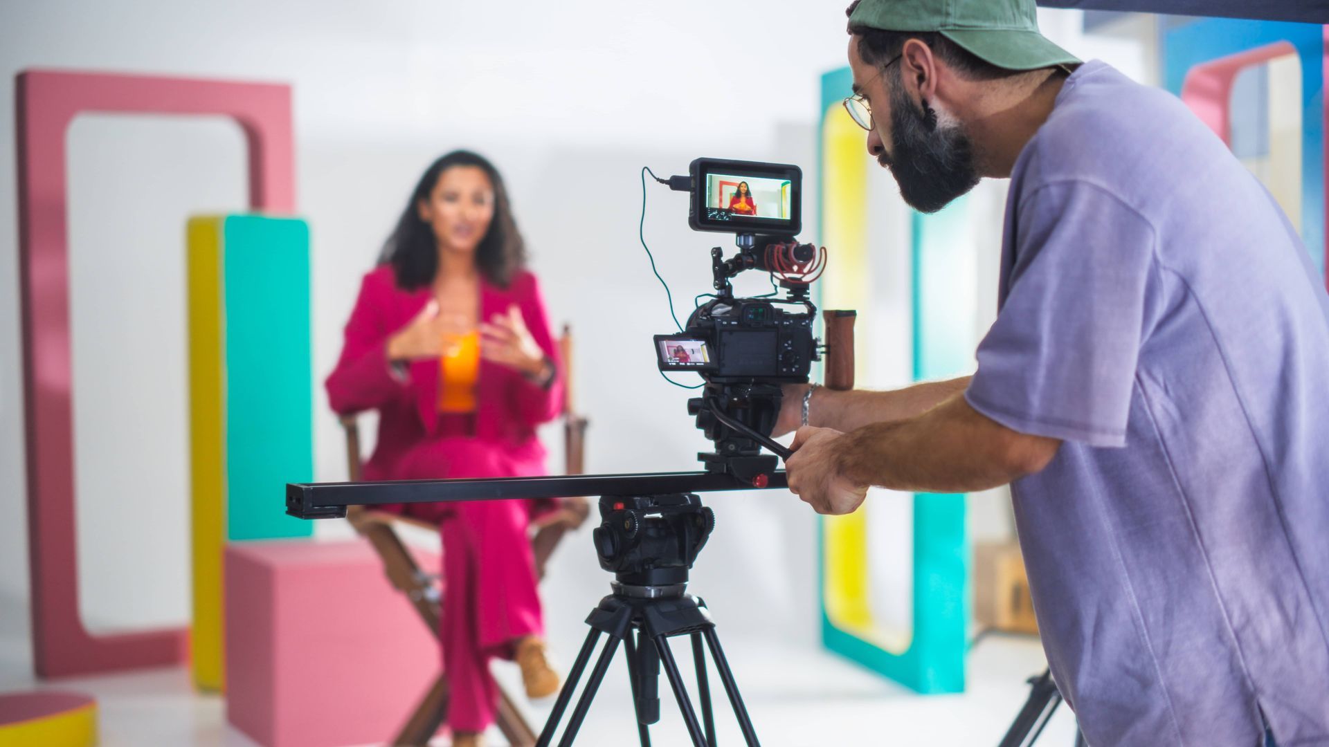 Filmmaker adjusts camera pointed at a woman in pink suit seated on set with colorful shapes.