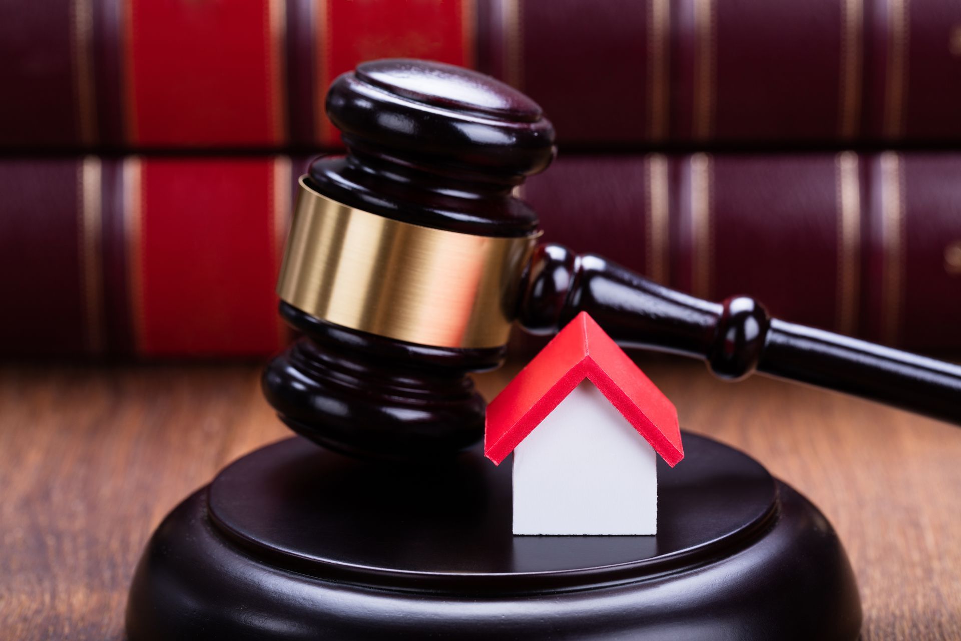 A gavel strikes a small house model on a wooden block, with law books in the background.