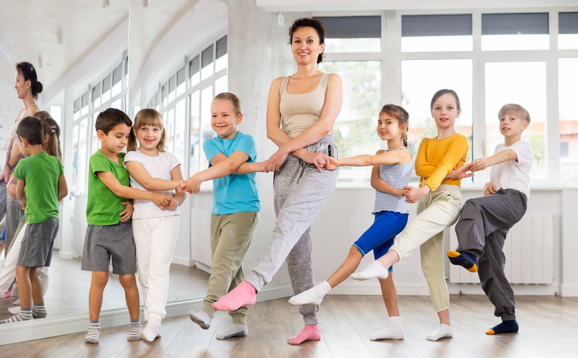 Instructor and children hold hands in a sunlit studio, each extending one leg outward in a stretch. Instructor and children hold hands in a sunlit studio, each extending one leg outward in a stretch.