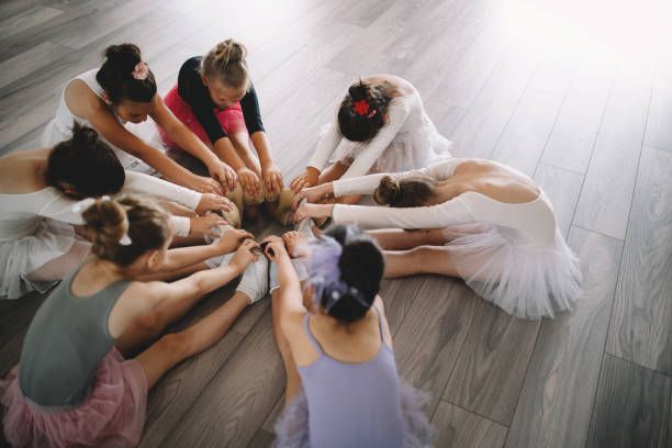 A group of ballet dancers in leotards and tutus sitting in a circle on a wooden floor, reaching toward the center.