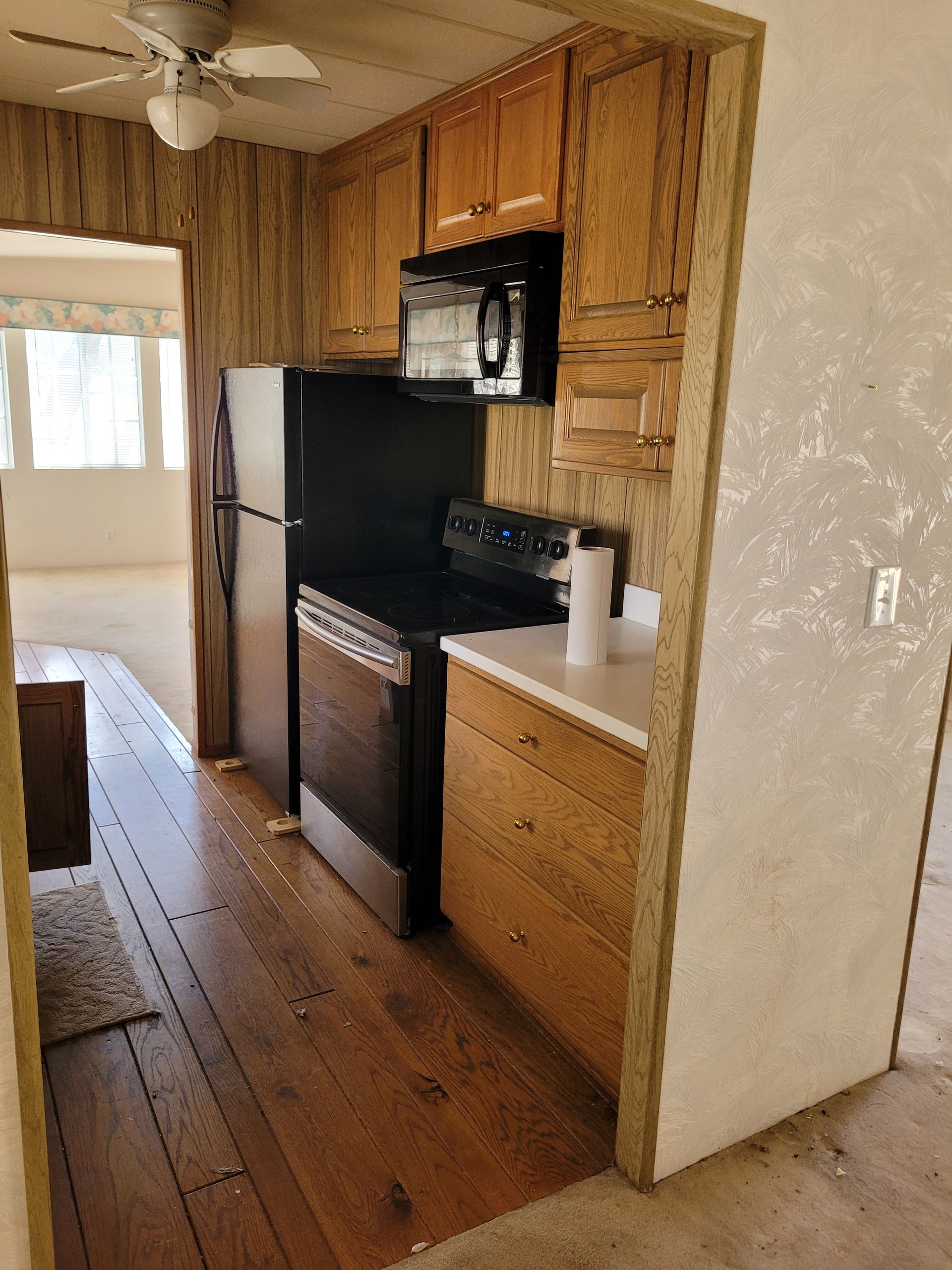 Narrow kitchen with wood cabinets, black appliances, and a view into the living room. The floor is wood.